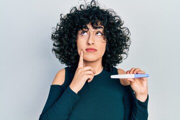 Young middle east woman holding pregnancy test result serious face thinking about question with...