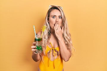 Beautiful young blonde woman wearing swimsuit drinking tropical cocktail covering mouth with hand,...