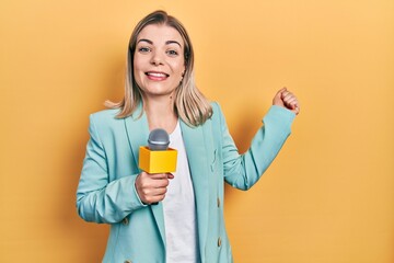 Beautiful caucasian woman holding reporter microphone screaming proud, celebrating victory and...