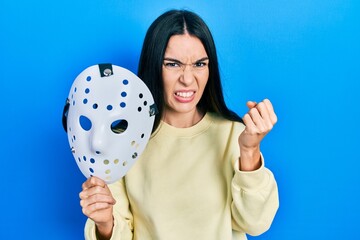 Young brunette woman holding hockey mask annoyed and frustrated shouting with anger, yelling crazy...