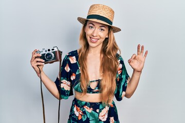 Young caucasian woman holding vintage camera doing ok sign with fingers, smiling friendly gesturing excellent symbol