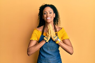 Middle age african american woman wearing professional apron praying with hands together asking for forgiveness smiling confident.