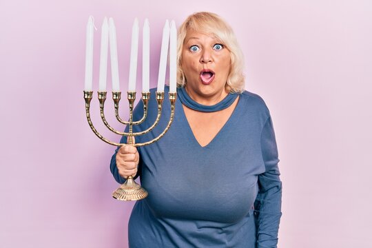 Middle age blonde woman holding menorah hanukkah jewish candle scared and amazed with open mouth for surprise, disbelief face