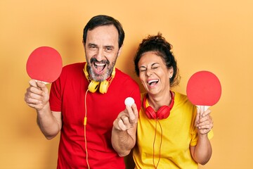 Middle age couple of hispanic woman and man holding red ping pong rackets smiling and laughing hard...