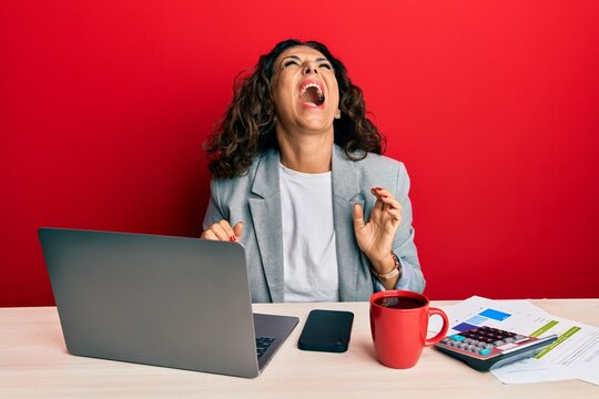 Beautiful middle age woman working at the office drinking a cup of coffee angry and mad screaming frustrated and furious, shouting with anger. rage and aggressive concept.