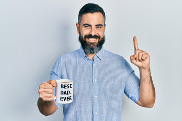 Young hispanic man drinking mug of coffee with best dad ever message smiling with an idea or question pointing finger with happy face, number one