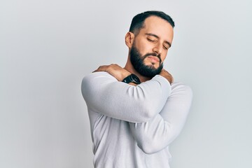 Young man with beard wearing casual white shirt hugging oneself happy and positive, smiling...