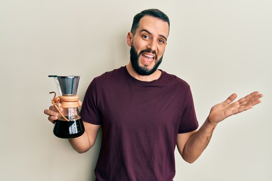 Young man with beard holding coffee maker with filter celebrating achievement with happy smile and winner expression with raised hand