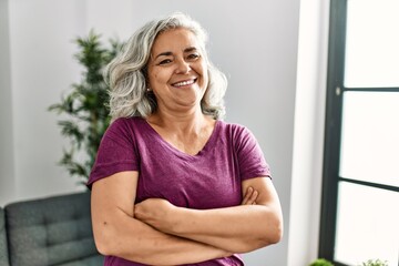 Middle age grey-haired woman smiling happy standing with arms crossed gesture at home.