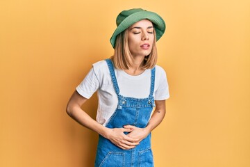 Young caucasian blonde woman wearing denim jumpsuit and hat with 90s style with hand on stomach...