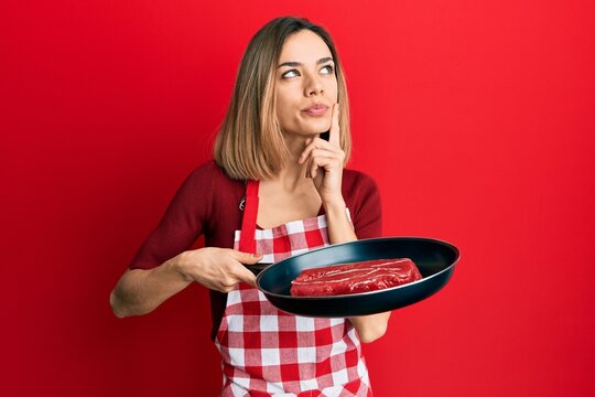 Young caucasian blonde woman wearing cook apron cooking meat on pan serious face thinking about question with hand on chin, thoughtful about confusing idea