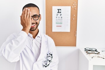 Fototapeta na wymiar African american optician man standing by eyesight test covering one eye with hand, confident smile on face and surprise emotion.
