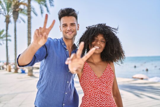Young interracial couple outdoors on a sunny day smiling looking to the camera showing fingers doing victory sign. number two.