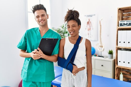 Physiotherapist working with patient wearing arm on sling at rehabilitation clinic winking looking at the camera with sexy expression, cheerful and happy face.