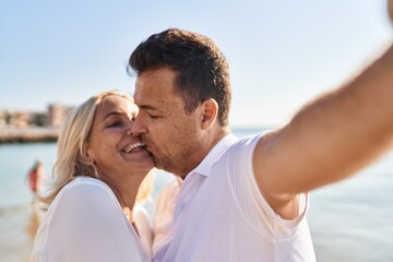 Fototapeta premium Middle age man and woman couple hugging each other and kissing make selfie by the smartphone at seaside
