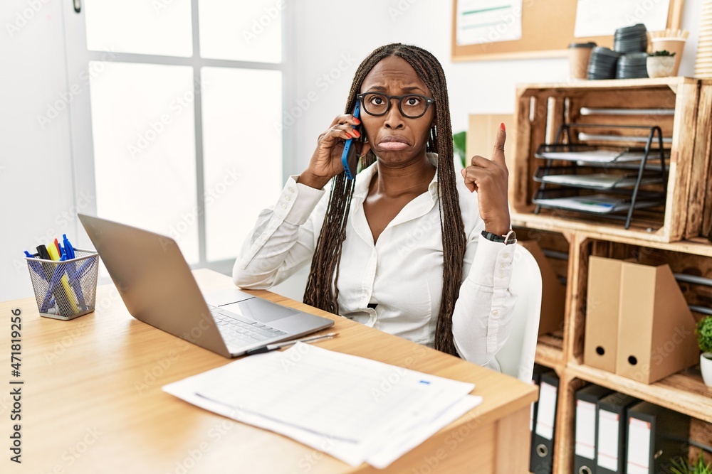 Poster Black woman with braids working at the office speaking on the phone pointing up looking sad and upset, indicating direction with fingers, unhappy and depressed.