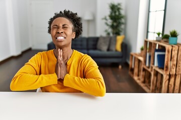 Young african american woman wearing casual clothes sitting on the table at home begging and...