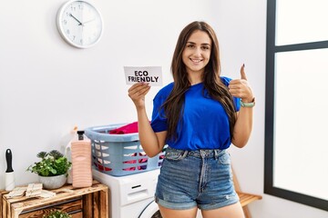 Young brunette woman doing laundry holding eco friendly paper smiling happy and positive, thumb up doing excellent and approval sign