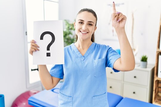 Young physiotherapist woman holding question mark surprised with an idea or question pointing finger with happy face, number one