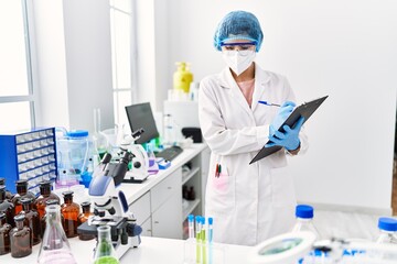 Young latin woman wearing scientist uniform writing on clipboard at laboratory