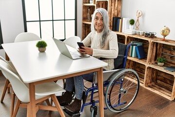 Middle age grey-haired woman using smartphone sitting on wheelchair at home