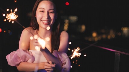 Young asian woman with sparklers is dancing and celebrating a new year. Fireworks, bengal lights in slow motion. Having fun at rooftop in the city.Happy interracial couple with glasses of wine .