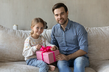 Portrait of smiling bonding young Caucasian father and little adorable child daughter sitting on sofa with wrapped gift box in hands, celebrating happy birthday or special occasion together at home.