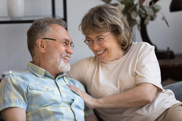 Happy sweet senior couple enjoying leisure time at home, having fun. Older grey haired husband and wife resting in living room, sitting on couch together, hugging with love, care, talking, laughing,