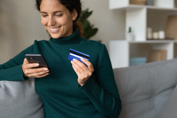 Web payment. Cropped shot of happy hispanic woman using cell for shopping paying online with bank card from home. Young female sit on couch make money transaction in convenient phone app. Copy space