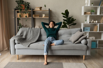 Obraz premium Sweet idleness. Lazy young hispanic lady sit in relaxed pose on big comfy sofa at living room interior breath air dream imagine. Successful female new flat apartment buyer rest at home feel pleasure