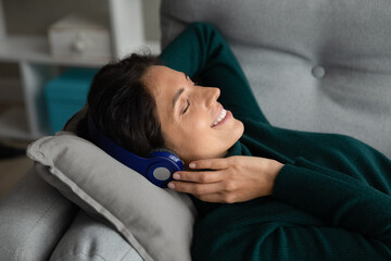 Melomane. Young hispanic woman music lover rest on sofa with earphones on head listen to chosen songs from playlist. Relaxed latin lady reduce stress with help of tranquil melodies in wireless headset