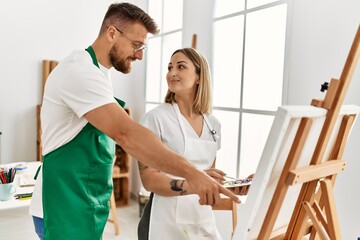 Fototapeta premium Young caucasian couple smiling happy drawing at art studio. Man standing and looking draw canvas.