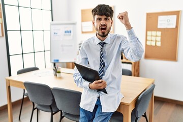 Hispanic man with beard wearing business style sitting on desk at office annoyed and frustrated shouting with anger, yelling crazy with anger and hand raised