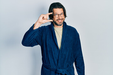 Middle age caucasian man wearing bathrobe and glasses smiling and confident gesturing with hand doing small size sign with fingers looking and the camera. measure concept.