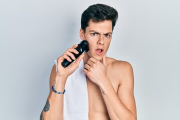 Young hispanic man shaving with electric razor machine in shock face, looking skeptical and sarcastic, surprised with open mouth