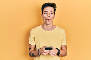 Young hispanic man using smartphone typing message making fish face with mouth and squinting eyes, crazy and comical.