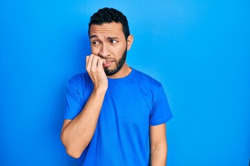 Hispanic man with beard wearing casual blue t shirt looking stressed and nervous with hands on mouth biting nails. anxiety problem.