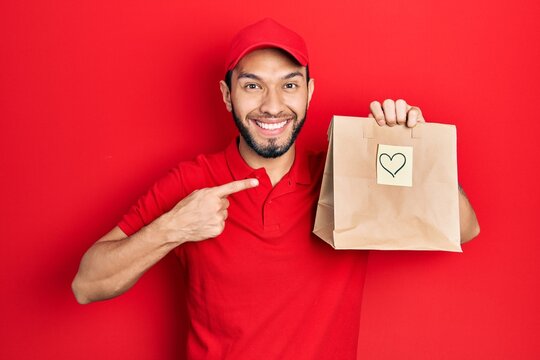 Hispanic man with beard holding take away paper bag with heart reminder smiling happy pointing with hand and finger