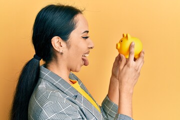 Young hispanic girl looking to piggy bank sticking tongue out happy with funny expression.