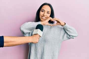 Young hispanic girl being interviewed by reporter holding microphone smiling in love doing heart...