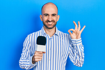 Naklejka premium Young bald man holding reporter microphone doing ok sign with fingers, smiling friendly gesturing excellent symbol