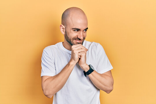 Young bald man wearing casual white t shirt laughing nervous and excited with hands on chin looking to the side