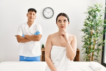 Young woman at wellness spa with professional therapist sitting on massage table serious face thinking about question with hand on chin, thoughtful about confusing idea