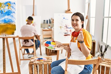 Young artist woman painting on canvas at art studio doing money gesture with hands, asking for salary payment, millionaire business
