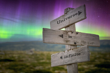 universe is infinite text quote on wooden signpost outdoors in nature with northern lights above.