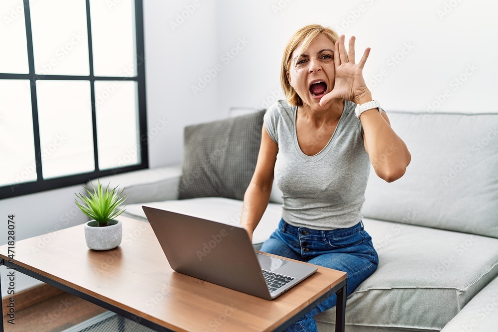 Poster middle age blonde woman using laptop at home shouting and screaming loud to side with hand on mouth.