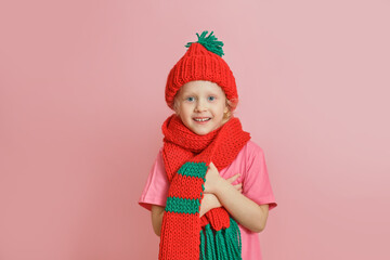caucasian toddler girl in red knitted scarf and hat on pink background, christmas mood concept copy space