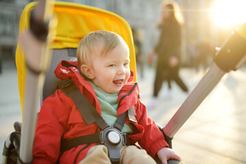 Sweet baby boy wearing red jacket sitting in a stroller outdoors. Little child in pram. Infant kid in pushchair. Spring walks with kids.