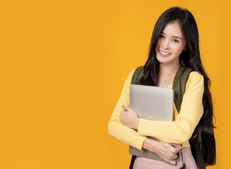 Happy young university student girl holding laptop computer with smile face carrying backpack Pretty asian woman stand over yellow background and copy space Beauty female look at camera with happiness