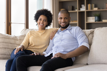 Happy millennial African American couple of homeowners, tenants head shot portrait. Husband and wife sitting on sofa, looking at camera, smiling, hugging with love, relaxing in new modern apartment